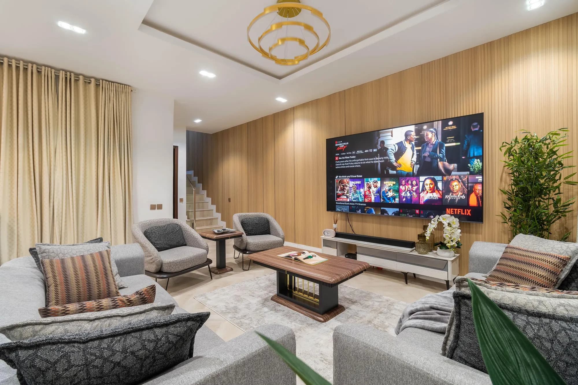 Living room with an oak-panelled TV wall, gold chandelier and a staircase beyond