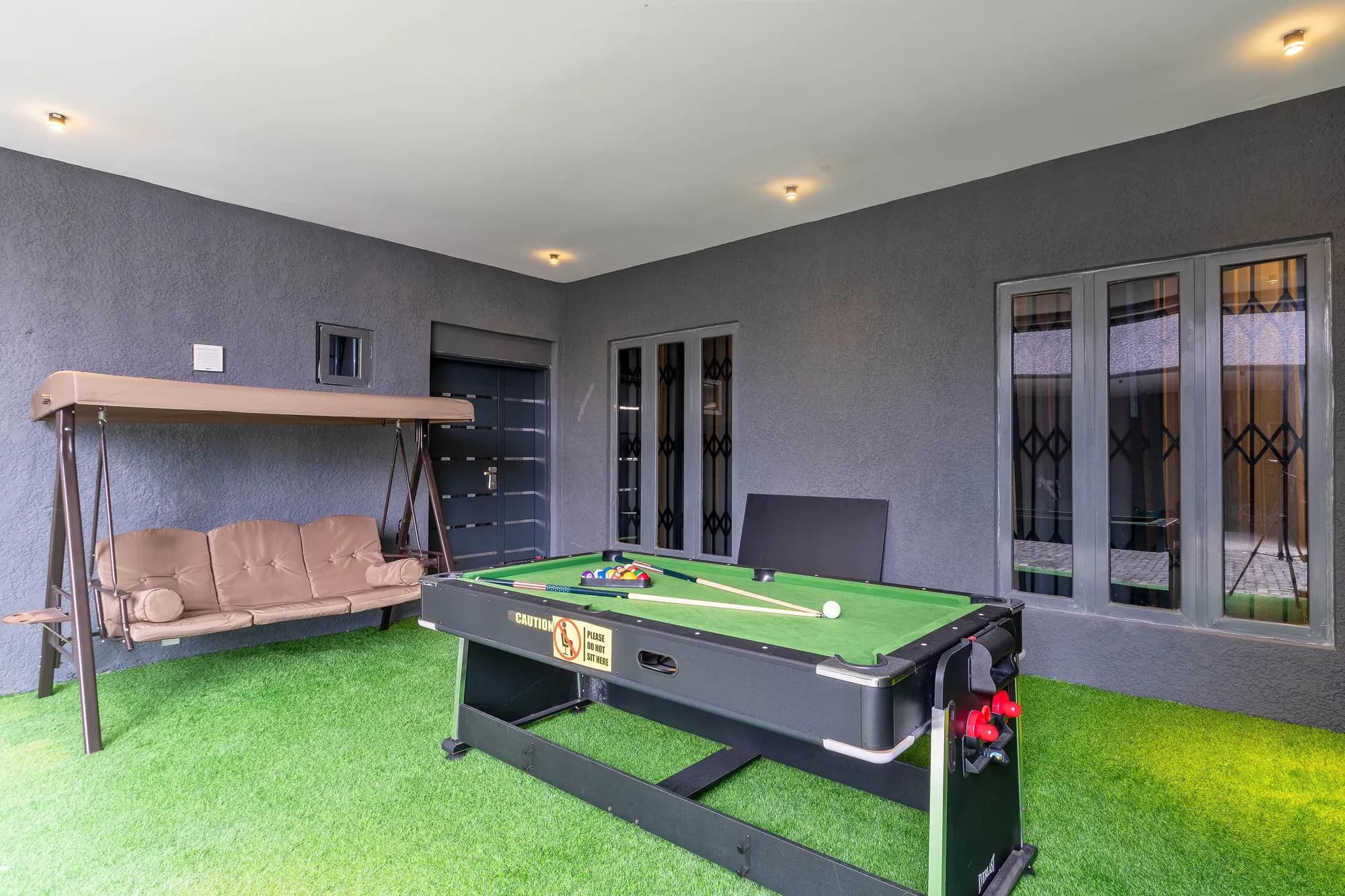 Covered games terrace with a green-baize pool table, cushioned garden swing and artificial turf